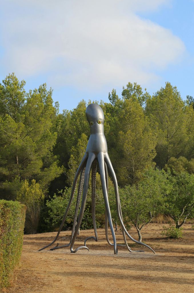 푸른 나무가 우거진 숲 앞에 서 있는 커다란 금속 문어 조각상.