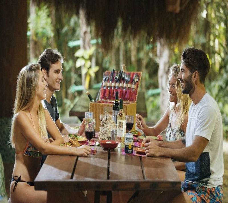 수영복을 입은 네 명이 나무 피크닉 테이블에 앉아 야외 열대 분위기에서 음식과 음료를 즐기고 있습니다.