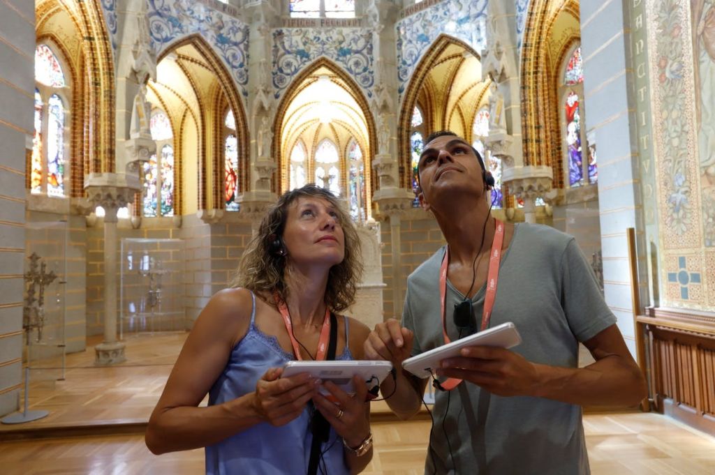 오디오 가이드를 착용한 두 사람이 아치형 천장과 스테인드글라스 창문이 있는 화려하고 대성당 같은 내부를 탐험합니다.