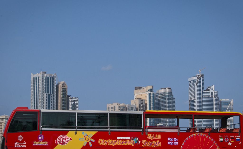 높은 현대식 건물과 맑고 푸른 하늘 앞에 '샤르자' 라고 표시된 레드 시티 관광 버스가 있습니다.