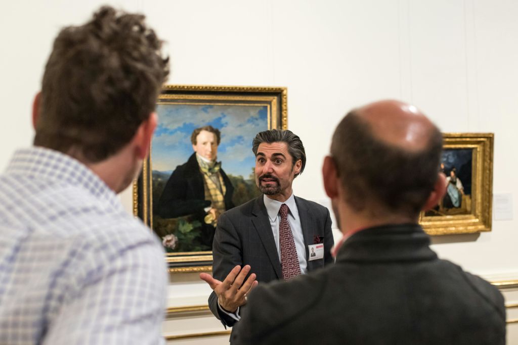 양복을 입은 한 남성이 역사적 인물의 초상화를 배경으로 두 남자와 대화하는 동안 몸짓을 하고 있습니다.