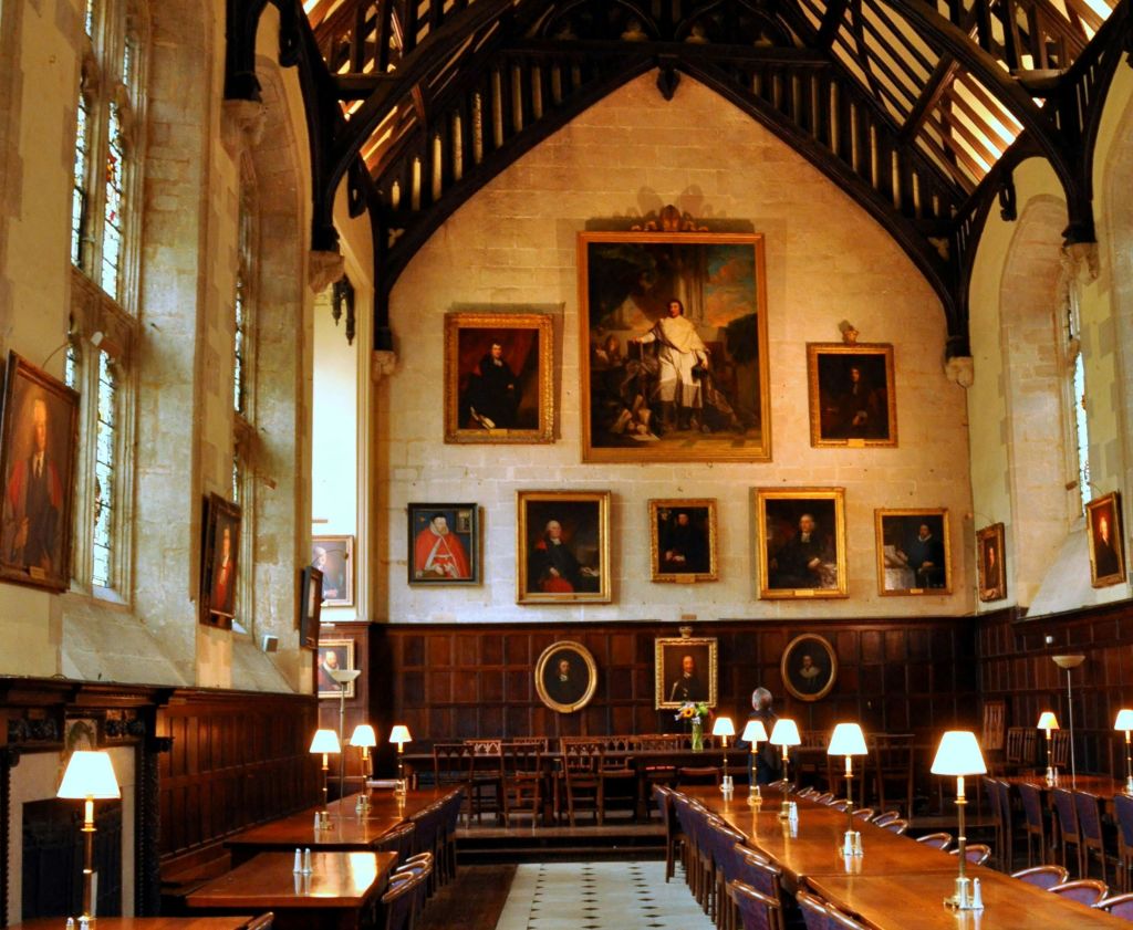 아치형 나무 대들보 천장 아래 긴 나무 테이블, 조명 램프, 초상화로 장식된 벽이 있는 유서 깊은 식당입니다.