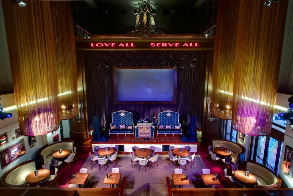 원형 테이블과 벨벳 의자, 대형 스크린, "LOVE ALL" 과 "SERVE ALL" 이라는 조명 표지판이 있는 세련된 라운지입니다.