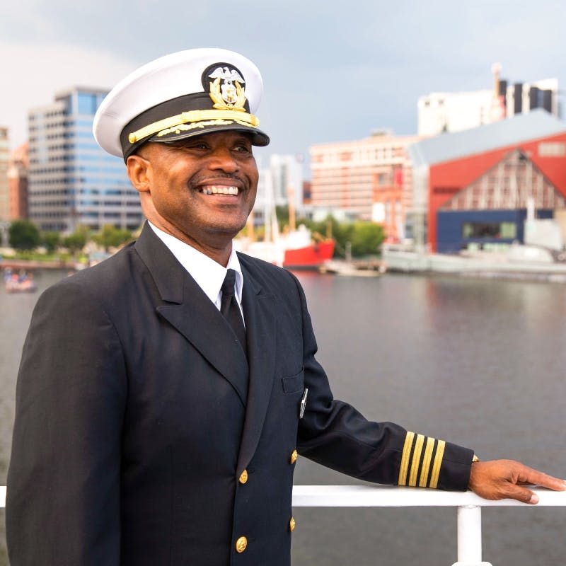 해군 제복을 입은 남성이 해안가 도시 풍경을 배경으로 배의 갑판에 서서 미소를 짓고 있습니다.