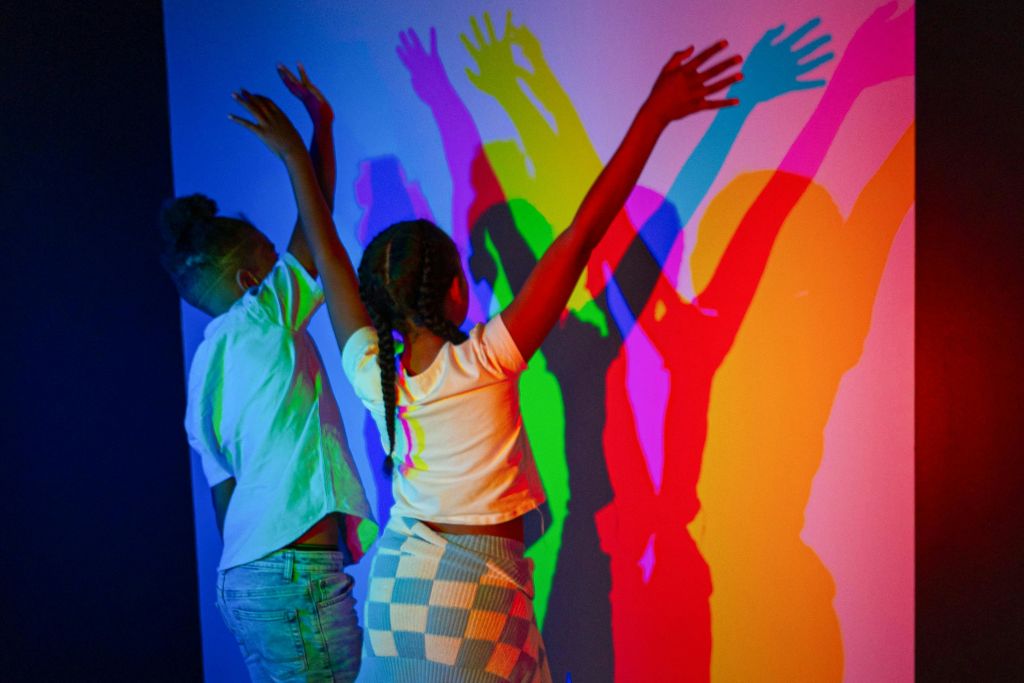 손을 들어 올린 두 아이가 여러 가지 빛깔의 조명 아래 벽에 겹쳐진 화려한 그림자를 드리웠습니다.