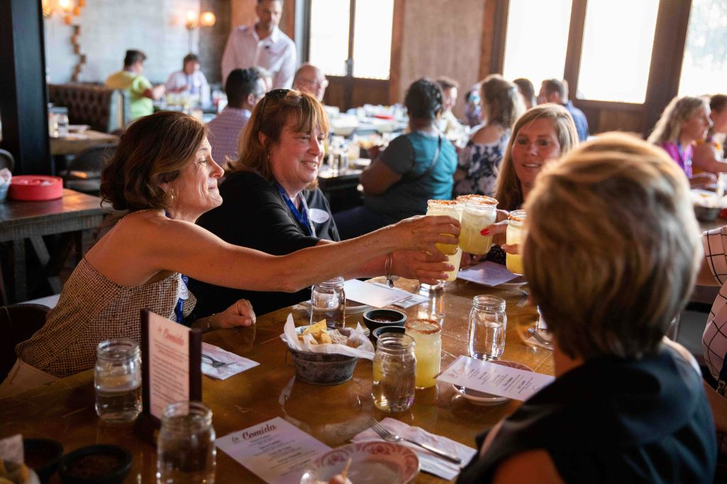 A group of people at a restaurant table toasting with drinks, with food and menus visible on the table.