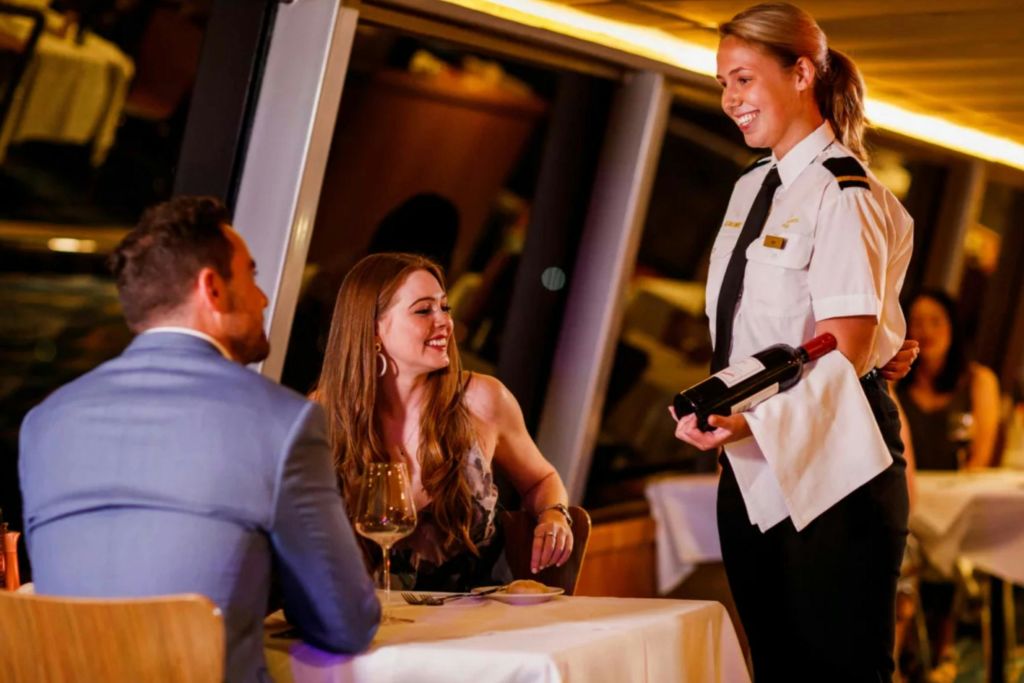 유니폼을 입은 웃는 웨이트리스가 와인병을 들고 테이블에 앉아 우아한 레스토랑을 바라보고 있습니다.