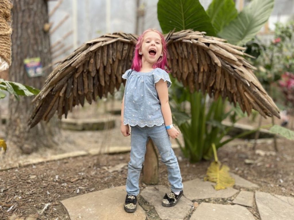 분홍색 머리를 한 어린 소녀가 나무 날개 구조물 앞에서 웃고 서 있는데, 날개가 있는 것처럼 보입니다. 밝은 파란색 옷을 입고 있습니다.