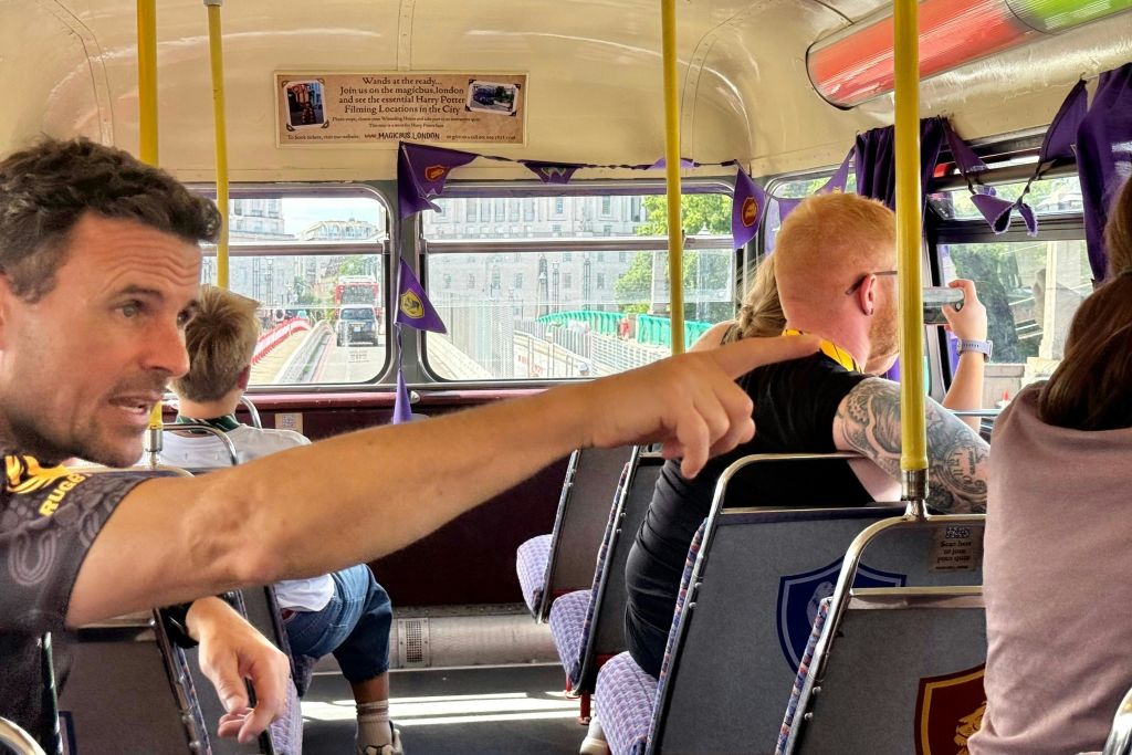 한 남성이 2층 버스 창문을 가리키고 있는데, 승객들이 앉아 있고 도시 풍경이 배경으로 보입니다.