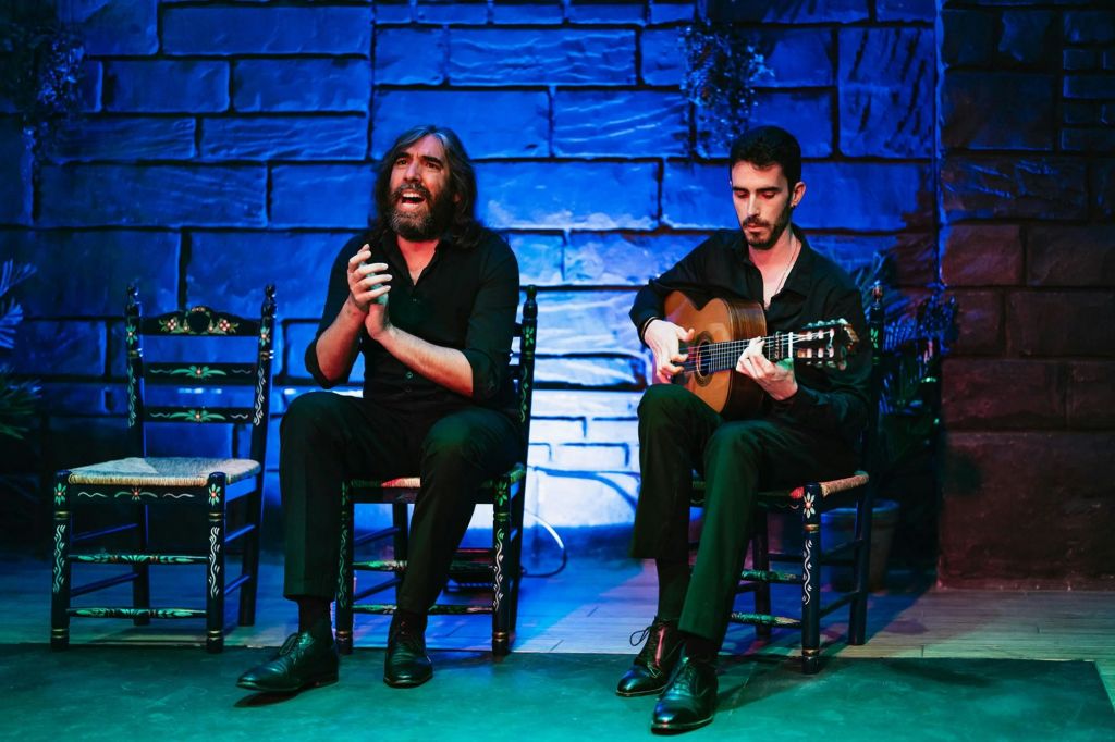 파란색 벽돌 벽 앞에서 기타를 연주하는 한 남자가 옆자리에 앉아 박수를 치며 노래하고 있습니다.