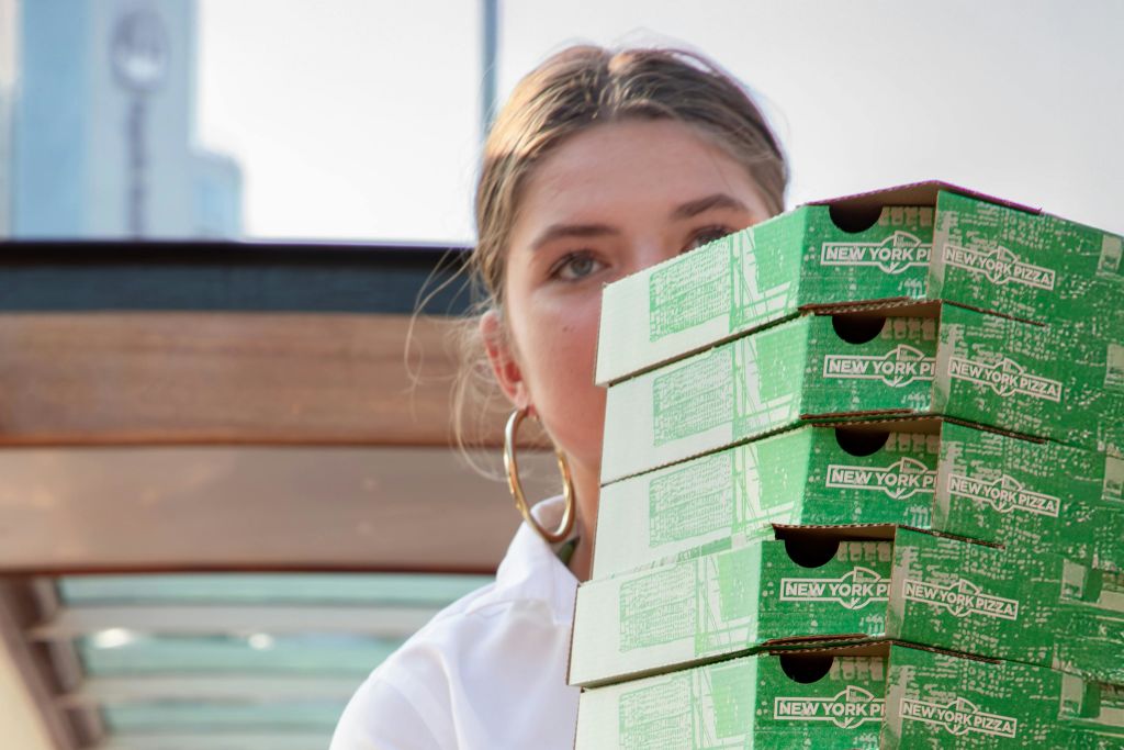 초록색 "뉴욕 피자" 상자를 들고 있는 사람으로, 코에서 위로 보이며, 실외 배경이 흐릿합니다.