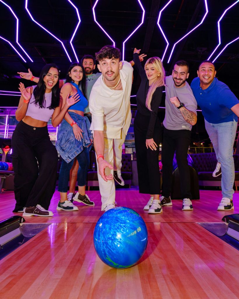 한 볼링장에서 7명의 친구들이 중앙에 있는 한 사람이 파란색 볼링공을 굴리고 있습니다. 위에 네온 불빛이 보입니다.