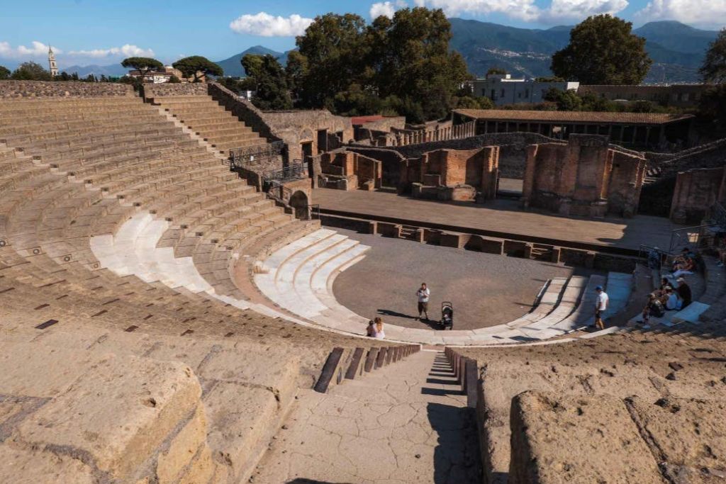 나무와 산으로 둘러싸인 계단식 석조 의자가 있는 고대 원형 극장. 아래에는 몇 명의 방문객이 있습니다.