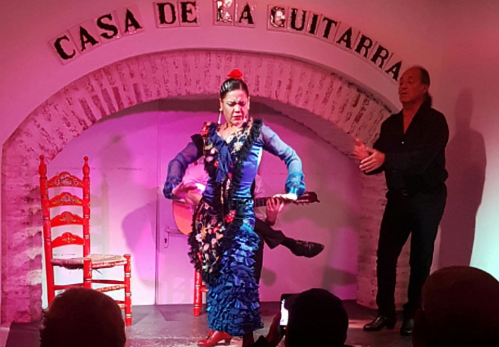 파란색 드레스를 입은 플라멩코 댄서가 "Casa de la Guitarra" 표지판이 있는 아치형 통로 아래 무대에서 기타리스트와 의자를 배경으로 공연하고 있습니다.