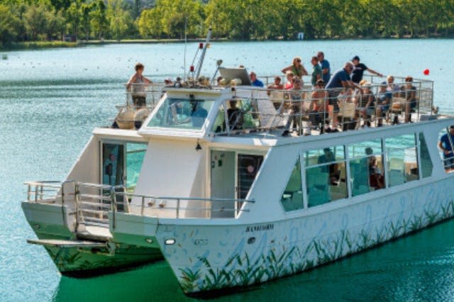 상부 갑판에 사람들이 있는 호숫가의 흰색 투어 보트. 나무와 푸른 해안선이 배경입니다.