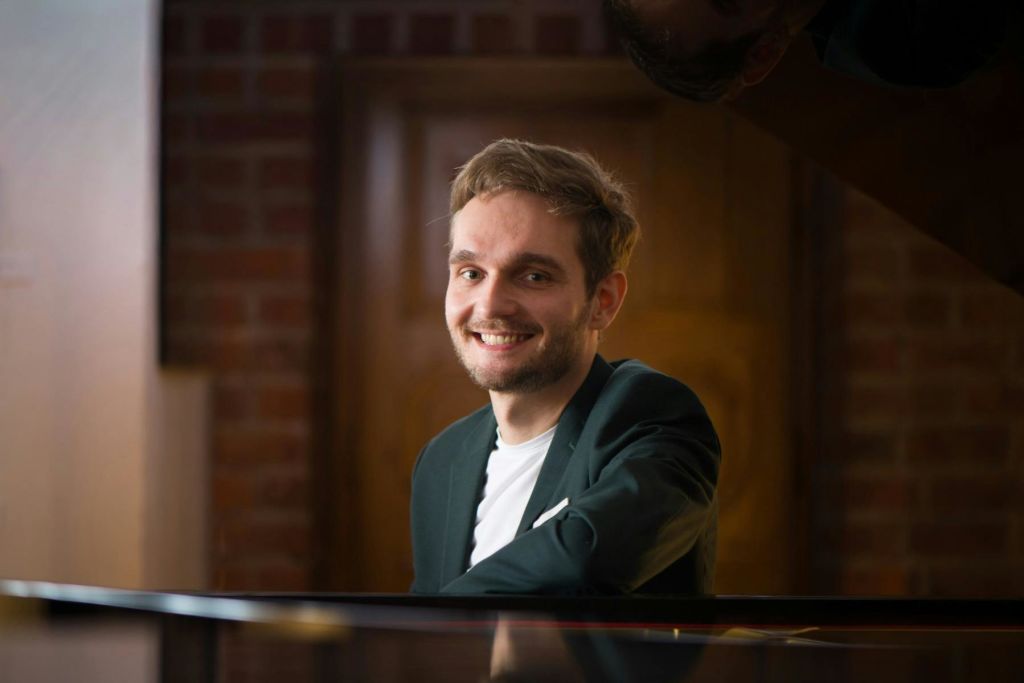 어두운 블레이저를 입은 웃는 사람이 흐릿한 나무 문과 벽돌 벽을 배경으로 피아노 근처에 앉아 있습니다.