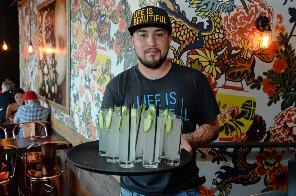 A man in a cap and T-shirt holding a tray of drinks in a colorful restaurant with patrons seated in the background.
