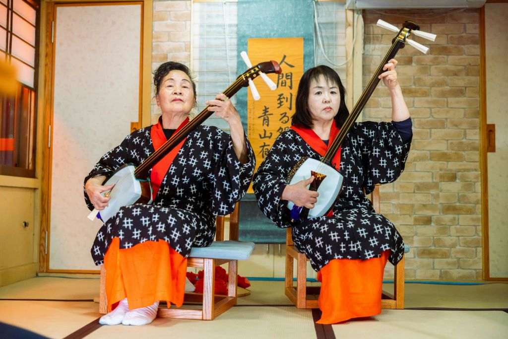 전통 복장을 한 두 여성이 벽돌 벽과 일본 서예가 있는 실내 공간에서 샤미센 악기를 연주합니다.