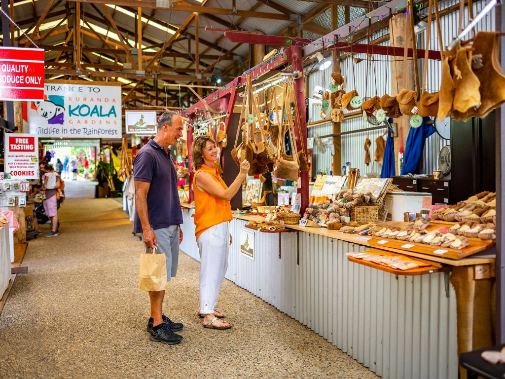 한 남녀가 '쿠란다 코알라 정원' 표지판 아래 시장 노점에서 수제품을 둘러보고 있습니다. 여자는 걸이식 물건을 살펴봅니다.