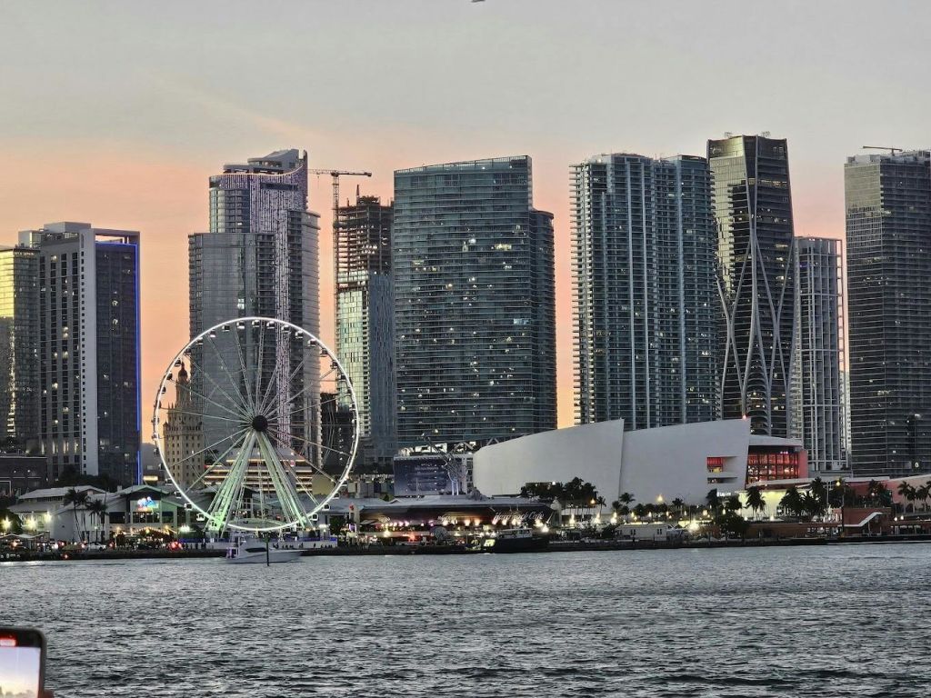 현대적인 고층 건물 근처에 조명이 켜진 대관람차가 있는 해질 무렵의 도시 스카이라인으로, 전경에 물이 흐릅니다.