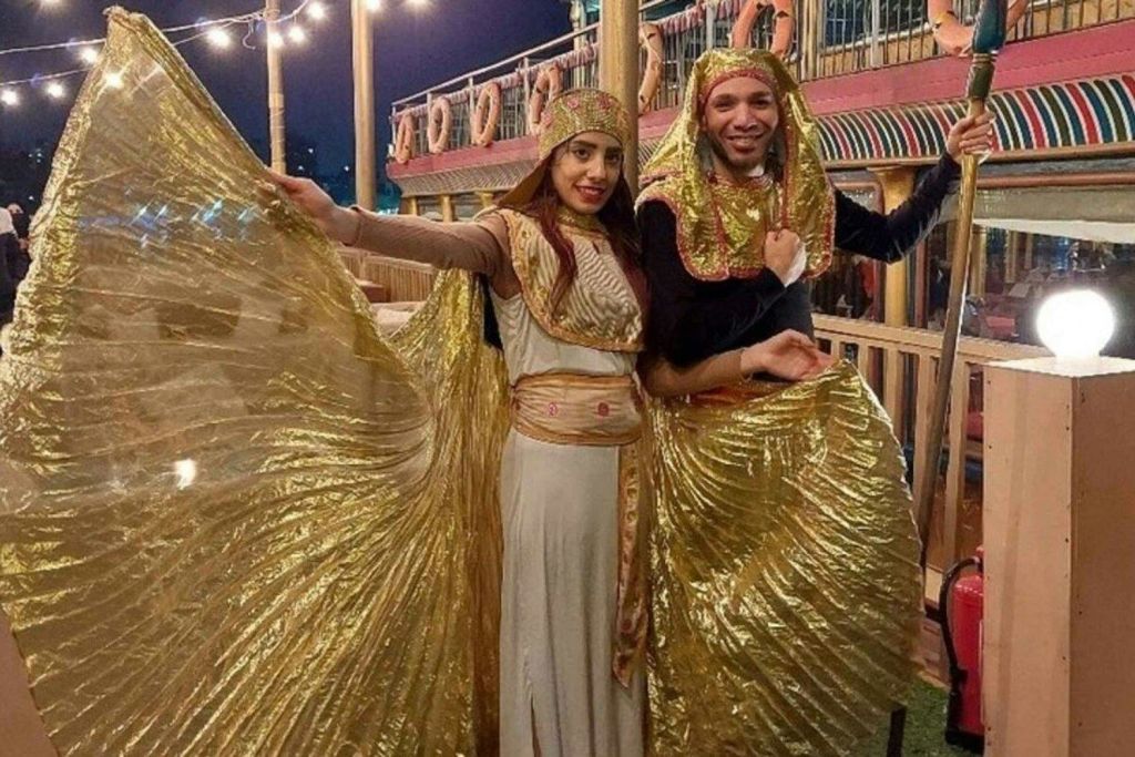 금색과 흰색의 이집트 스타일의 의상을 입은 두 사람이 생명 반지와 현악기가 달린 갑판형 보트에서 포즈를 취하고 있습니다.