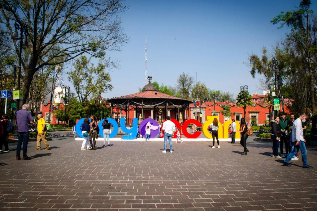 '코요아칸 (Coyoacán)' 이라는 알록달록한 철자가 새겨진 공공 광장, 산책하는 사람들, 나무로 둘러싸인 정자.