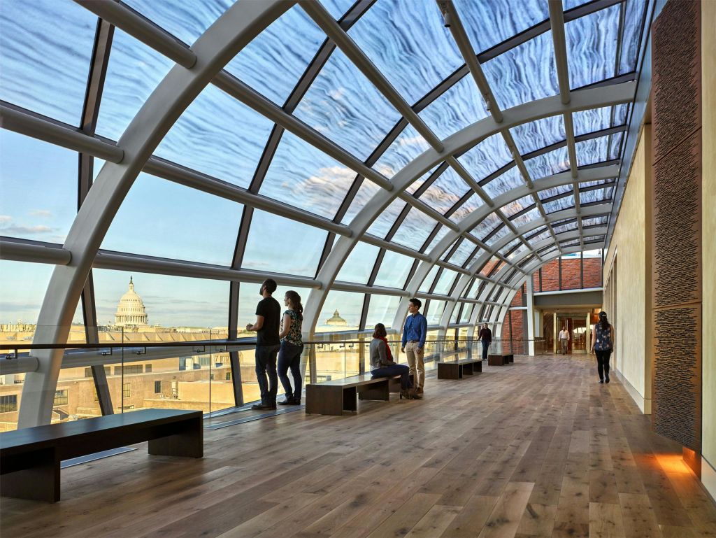 사람들은 곡선 지붕이 있는 통유리로 둘러싸인 복도에 서서 멀리 돔형 건물이 있는 도시 풍경을 감상합니다.