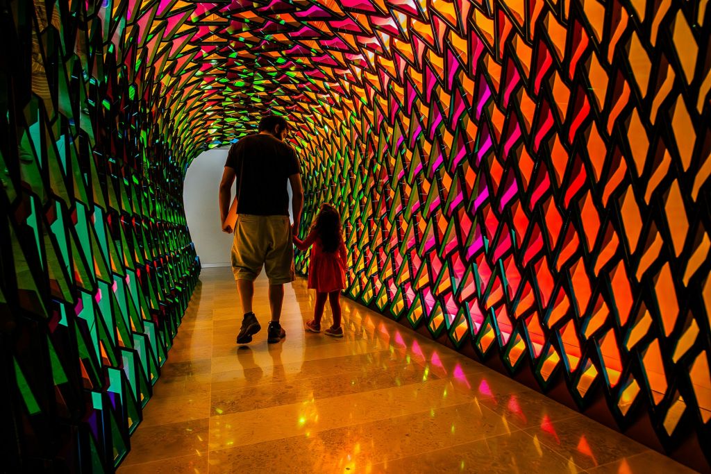 한 남자와 어린이가 다채롭고 반사되는 기하학적 벽이 있는 터널을 거닐며 생동감 넘치는 빛의 무늬로 조명을 받습니다.