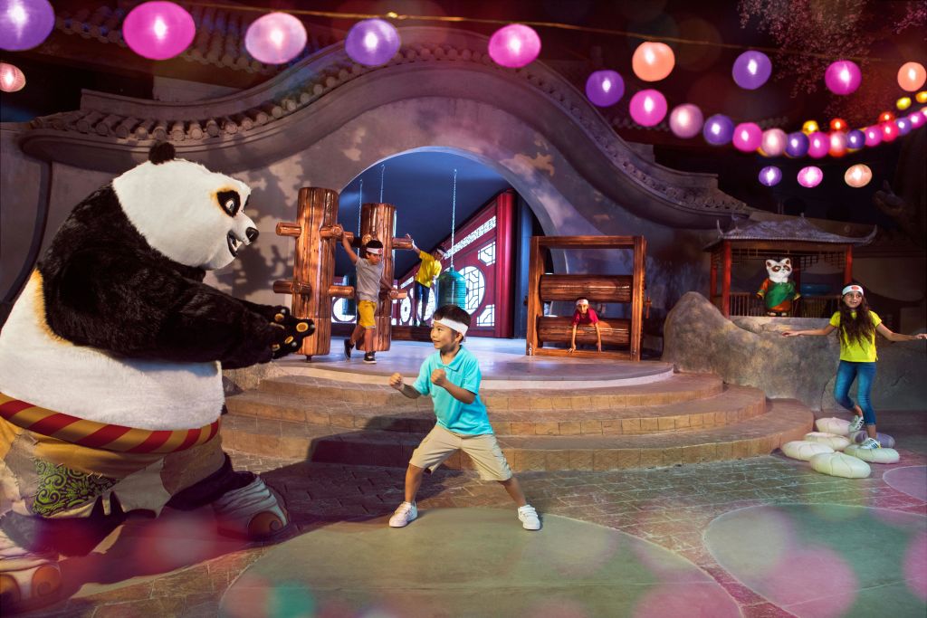 한 어린이가 테마가 있는 환경에서 커다란 판다 마스코트와 장난스럽게 포즈를 취하고 있으며, 위에 화려한 등불이 걸려 있습니다.