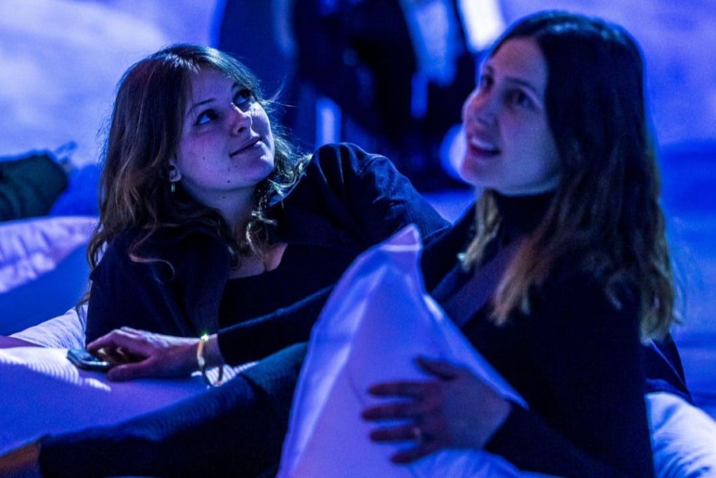 파란색 조명이 있는 조명이 희미한 방에 있는 두 명의 여성이 한 명은 베개를 들고 있으며, 둘 다 편안하고 약혼한 것처럼 보입니다.