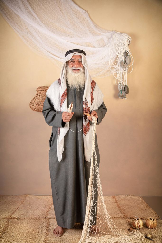 전통적인 복장을 한 긴 흰 수염을 가진 노인이 드레이프 그물로 베이지색 배경에 어망 수리 도구를 들고 있습니다.
