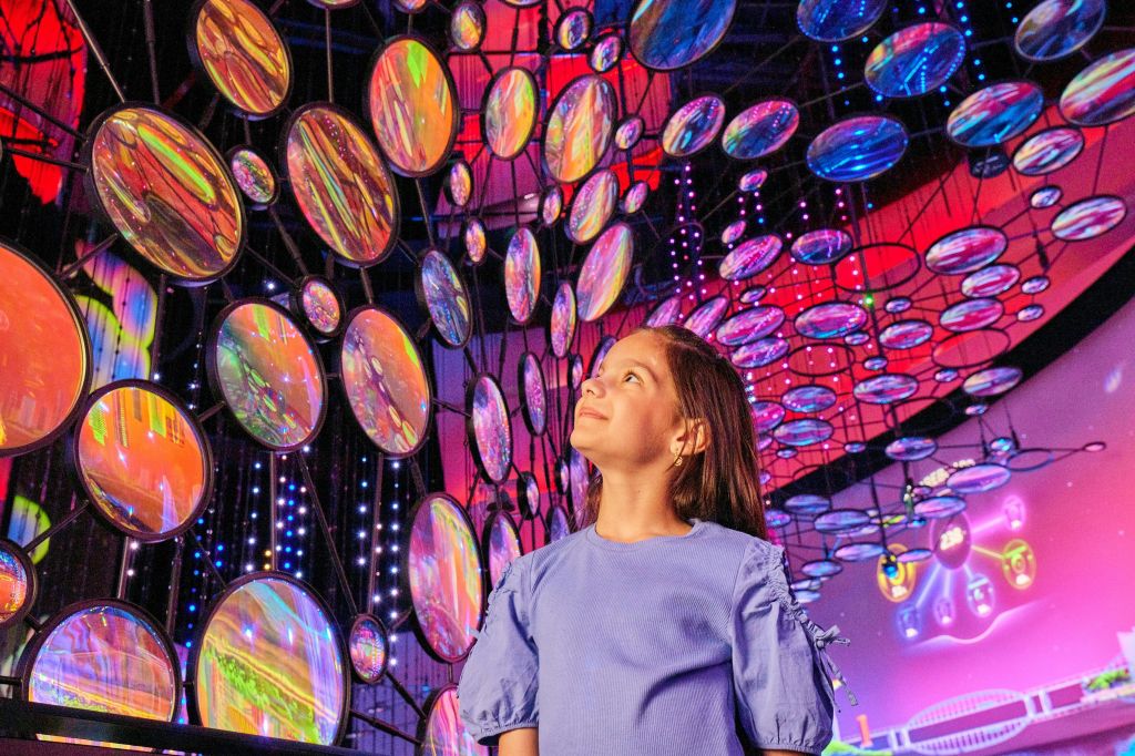파란색 드레스를 입은 한 소녀가 생동감 있고 예술적인 분위기에서 화려하고 반사되는 원형 패널과 조명을 올려다보고 있습니다.
