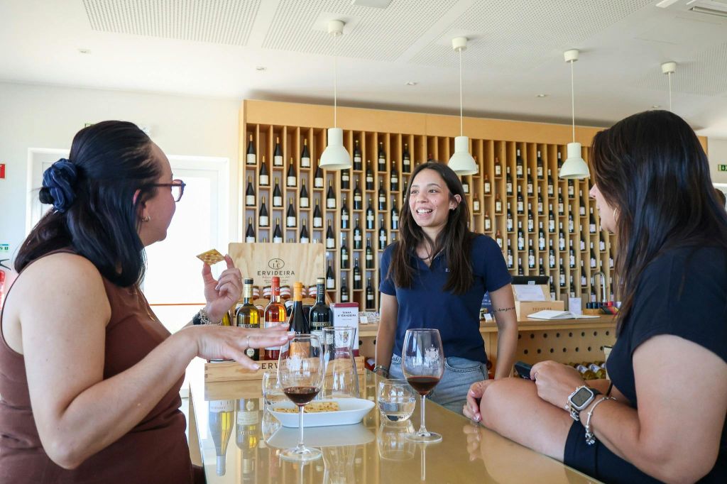 와인 랙 앞 카운터에서 와인 시음을 즐기는 세 사람. 잔과 병이 모두 보입니다.