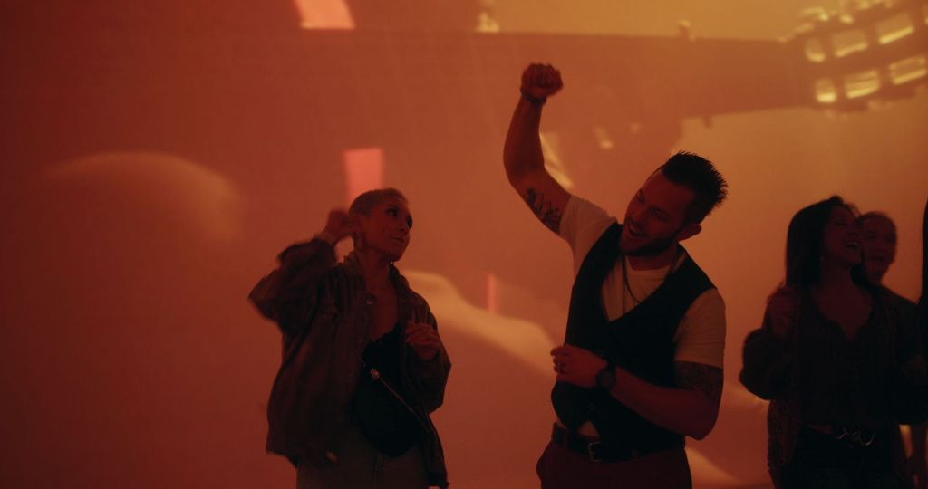 따뜻한 톤의 추상적인 배경에 열정적인 표정으로 춤을 추는 두 사람. 한 명은 주먹을 들어 올리고, 다른 한 명은 위를 바라봅니다.