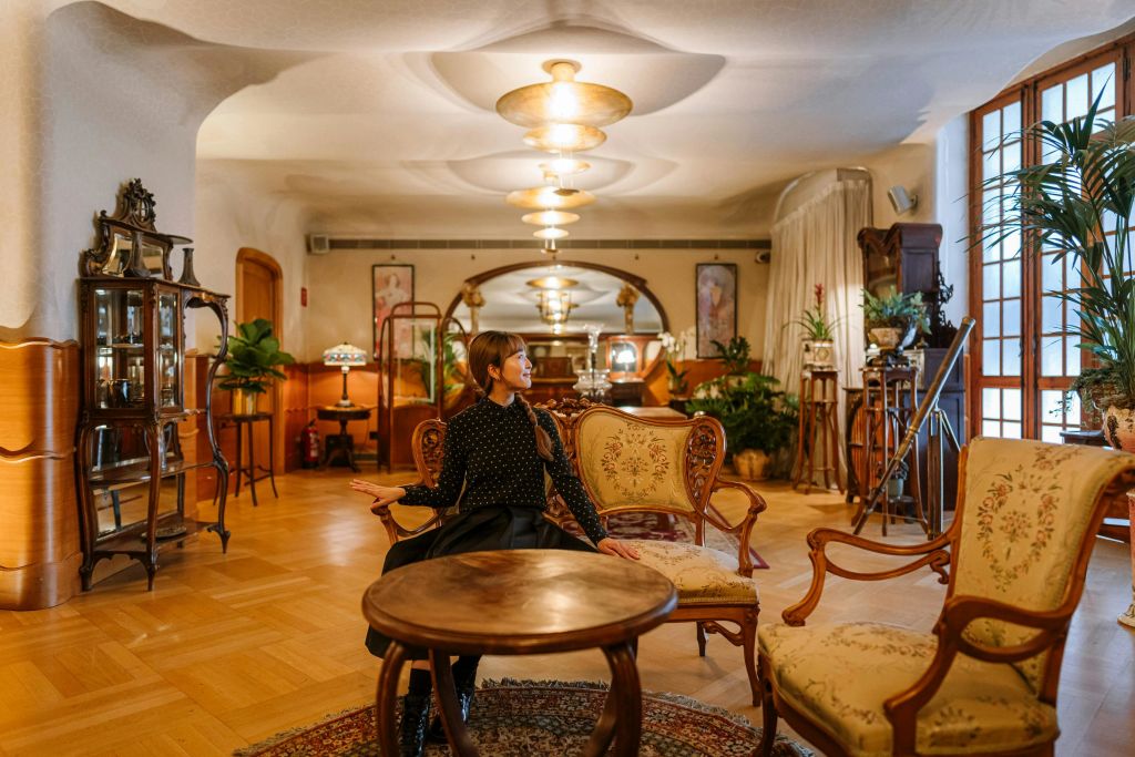 빈티지한 인테리어와 목재 가구, 따뜻한 조명이 돋보이는 우아한 객실에서 한 사람이 화려한 안락의자에 앉아 있습니다.