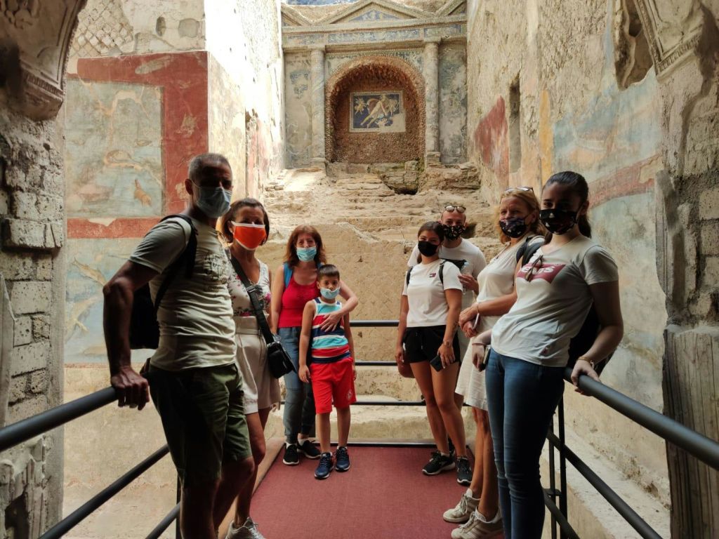 석조 계단을 배경으로 벽화로 장식된 고풍스러운 방에 가면을 쓴 사람들이 플랫폼에 서 있습니다.