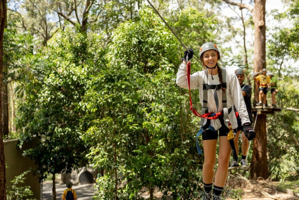 안전 장비를 착용한 사람이 숲을 가로지르며 미소를 짓고, 다른 사람들은 플랫폼을 배경으로 기다립니다.