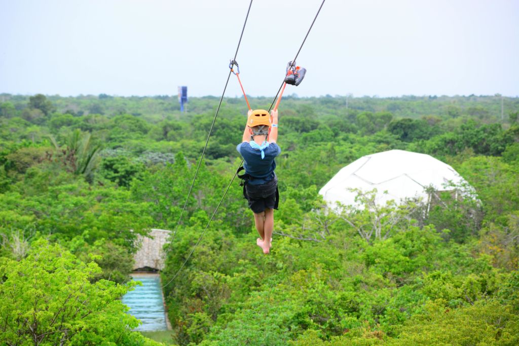 무성한 녹색 풍경 위에 사람이 지퍼 라이닝을 하고, 헬멧과 하네스를 착용하고 있으며, 배경에 흰색 돔 구조가 보입니다.