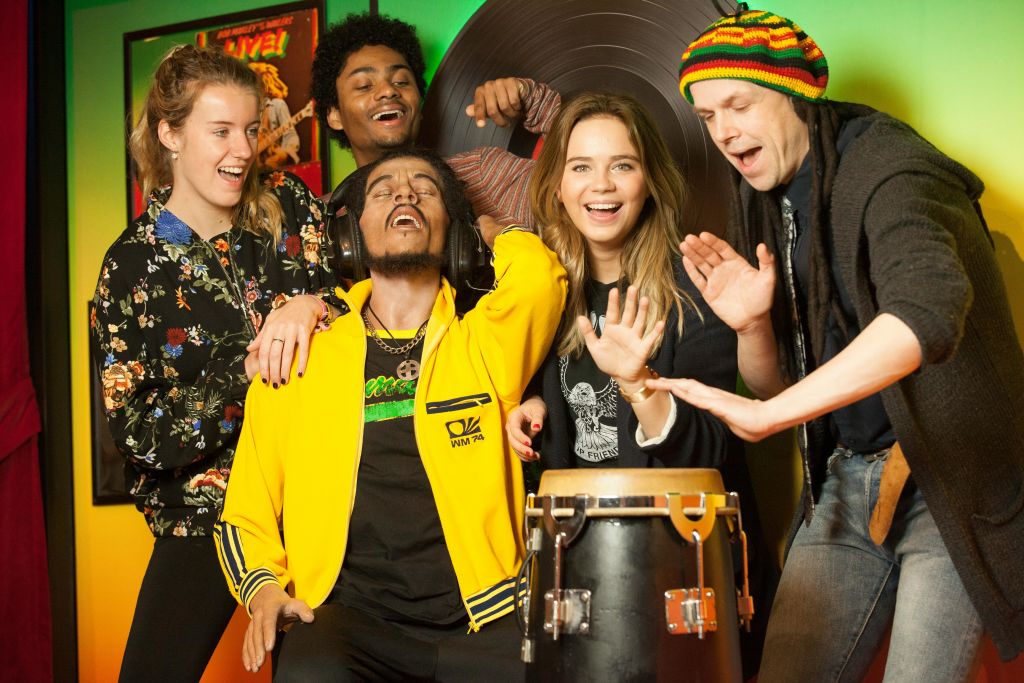 다섯 명이 실내에서 함께 음악을 즐기고, 한 명은 드럼을 연주하고, 다른 한 명은 헤드폰을 씁니다. 그들은 활기차고 즐겁게 보입니다.