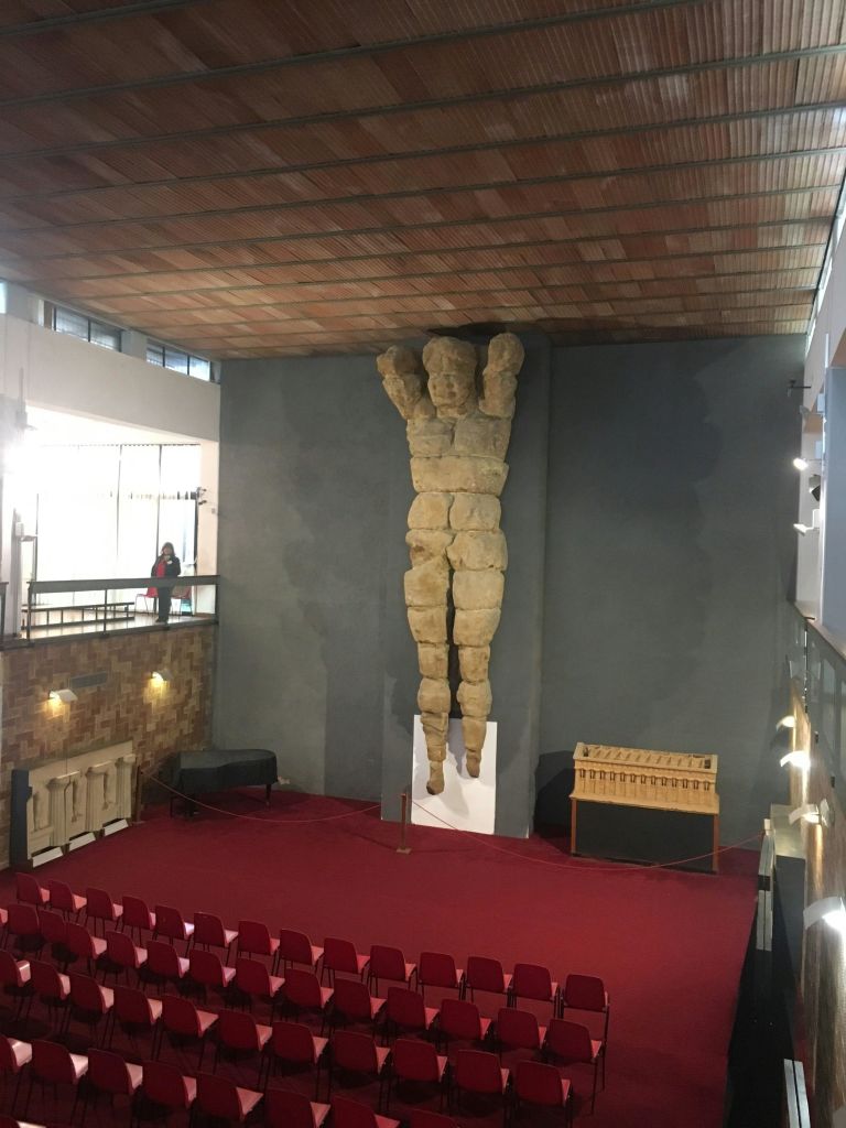 벽에 박힌 커다란 석상, 양팔을 들어 올리고 나무 천장이 있는 넓은 객실에서 붉은 의자가 줄지어 내려다보입니다.