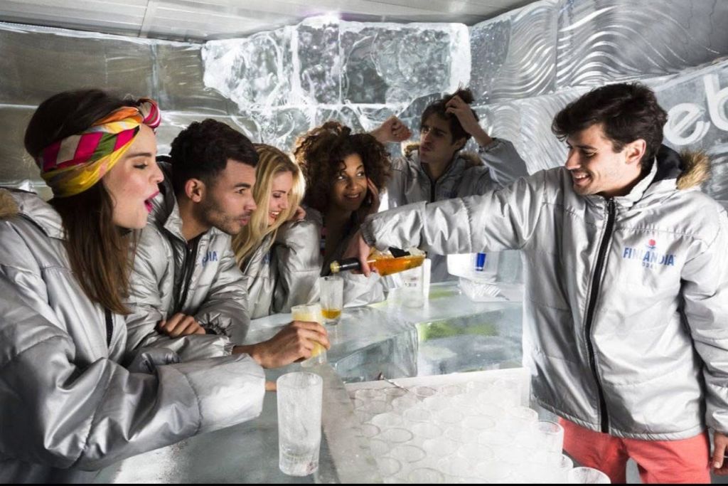 실버 재킷을 입은 한 무리의 사람들이 벽과 카운터에 얼음 장식이 있는 바에서 음료를 즐기고 있습니다.