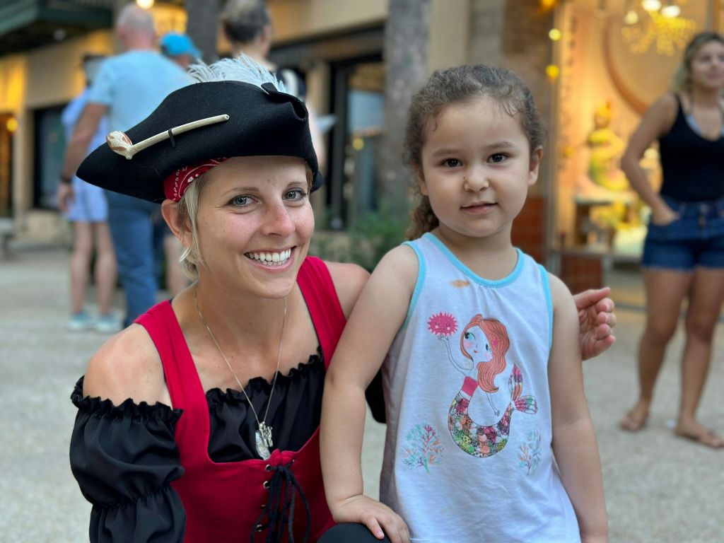 해적 의상을 입은 여성이 인어공주를 테마로 한 셔츠를 입은 어린 소녀 옆에서 미소를 짓고 있습니다. 상점 근처의 야외에 있습니다.