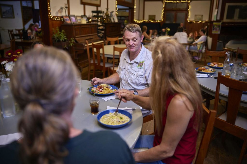 세 사람이 아늑한 레스토랑의 원형 테이블에 앉아 파스타를 먹고 있습니다. 다른 손님은 백그라운드에서 볼 수 있습니다.