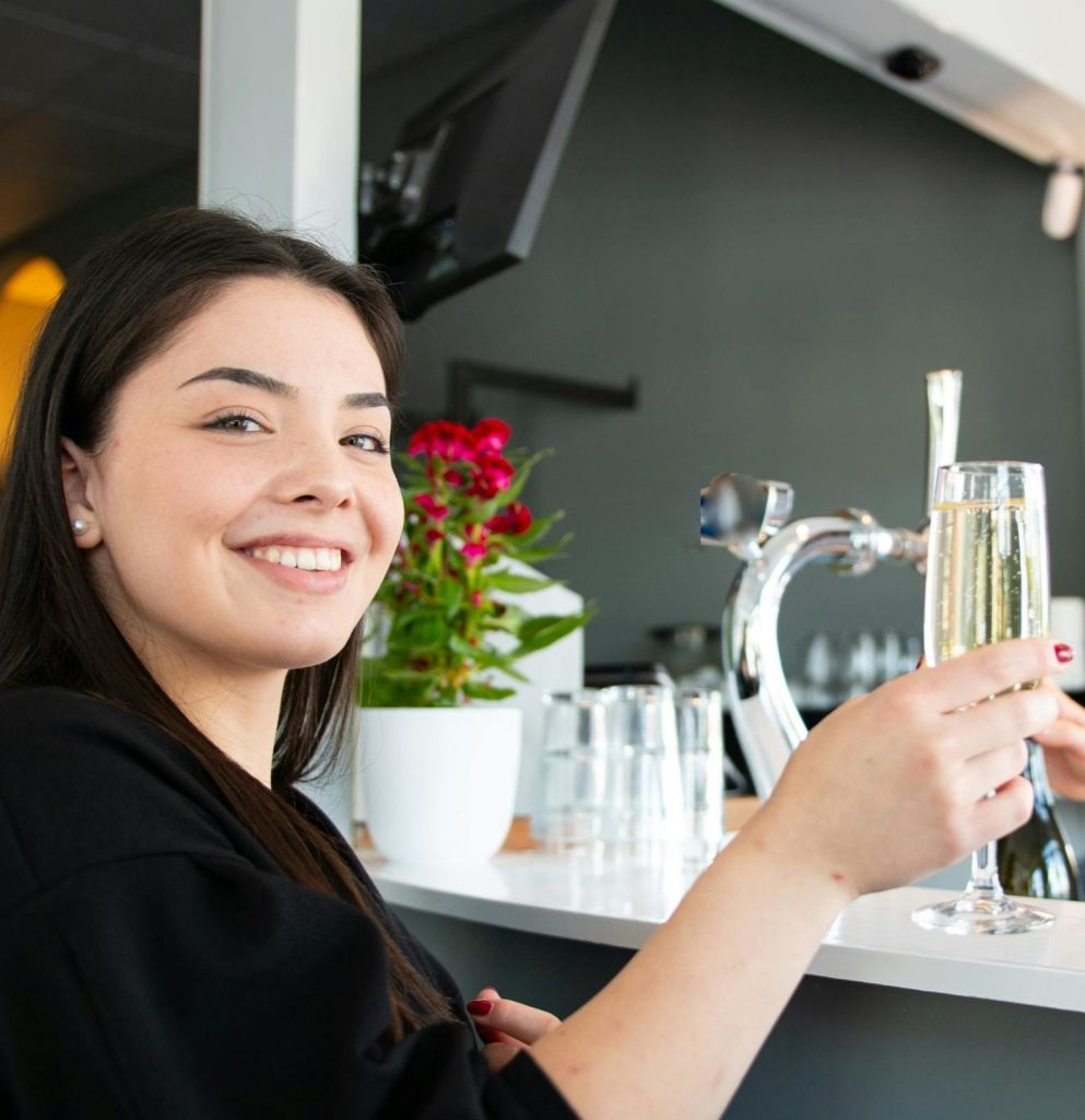 식물과 바 도구를 배경으로 한 바에서 샴페인 한 잔을 들고 미소를 짓는 여성.