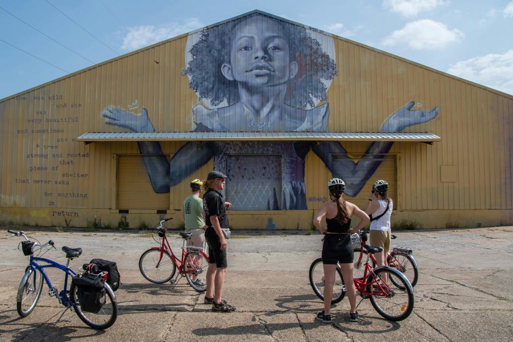 팔을 쭉 뻗고 곱슬머리를 한 사람의 커다란 벽화가 있는 건물 앞에 한 무리의 자전거 타는 사람들이 서 있습니다.