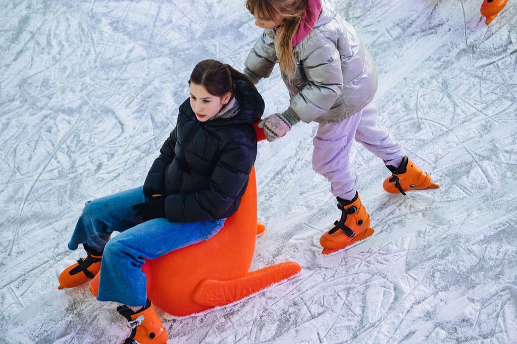 두 사람이 아이스링크를 타고 있습니다. 한 명은 주황색 얼음 썰매에 앉아 있고, 다른 한 명은 뒤에서 밀고 있습니다. 둘 다 겨울 의류와 아이스 스케이트를 착용합니다.