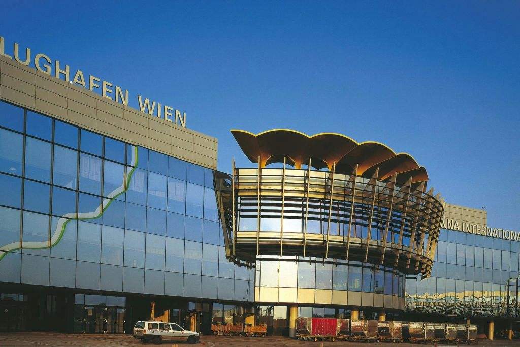 "FLUGHAFEN WIEN" 이라는 문구가 있는 현대적인 건물의 유리 외관과 독특한 건축 구조, 차량과 수하물 카트가 주차되어 있습니다.