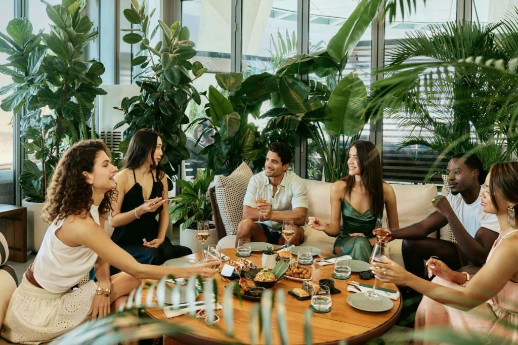 조명이 밝은 방의 울창한 실내 식물로 둘러싸인 테이블에 네 명이 앉아 음료와 간식을 즐기고 있습니다.