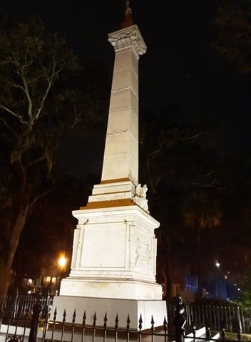 밤에는 나무와 조명으로 둘러싸인 공원에 정교한 조각이 있는 높고 밝은 흰색 기념비가 서 있습니다.