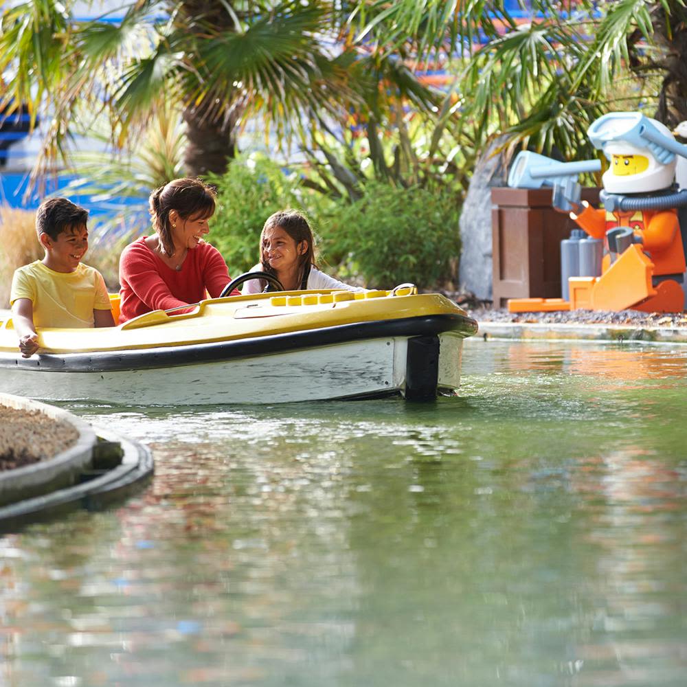 근처에 파란색과 주황색 로봇 동상이 있는 야자수로 둘러싸인 노란색 보트를 탄 세 명의 사람이 수상 자전거를 타고 있습니다.